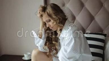 早晨，金发姑娘穿着白色衬衫坐在窗边的床上